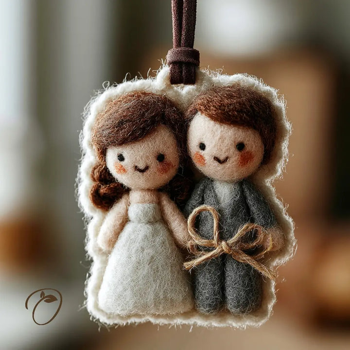 A felt wool hanging ornament depicting a cute bride and groom couple, with a brown hanging loop at the top. The bride is made of cream-colored wool with brown hair, wearing a white dress.