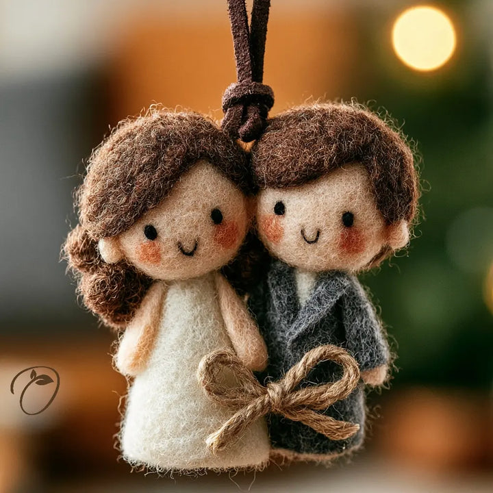 A felt wool hanging ornament shaped like a couple embracing, with a dark brown hanging cord at the top. The woman figure wears an off-white dress, and the man figure wears a dark gray robe.