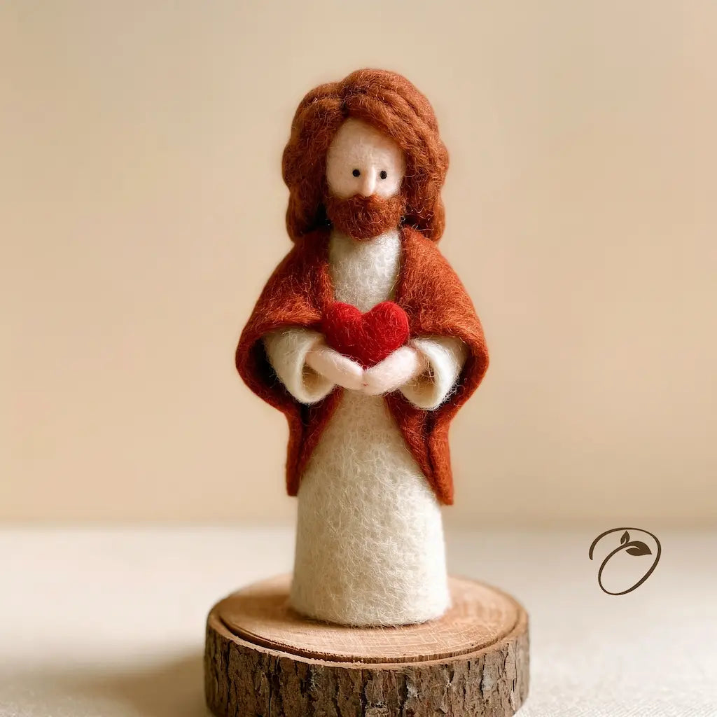 A handcrafted felt figurine of Jesus Christ standing on a small wooden slice base, holding a red heart. The figure has brown wool hair and beard, a white robe, and a rust-brown cloak, set against a soft beige background.