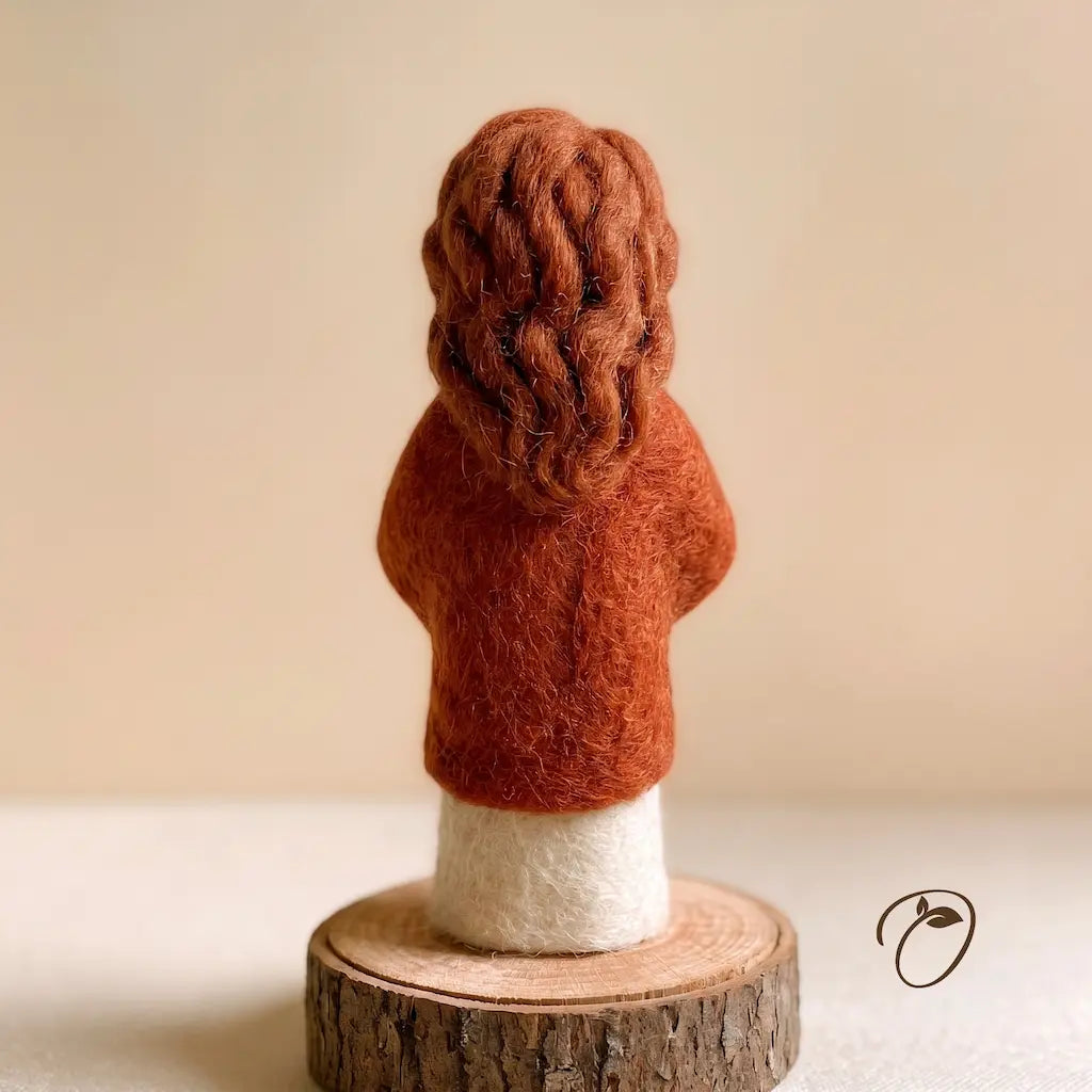 A handcrafted felted wool figurine of a child seen from the back, wearing a rust-orange sweater and cream pants, standing on a rustic wood slice base against a warm beige background.