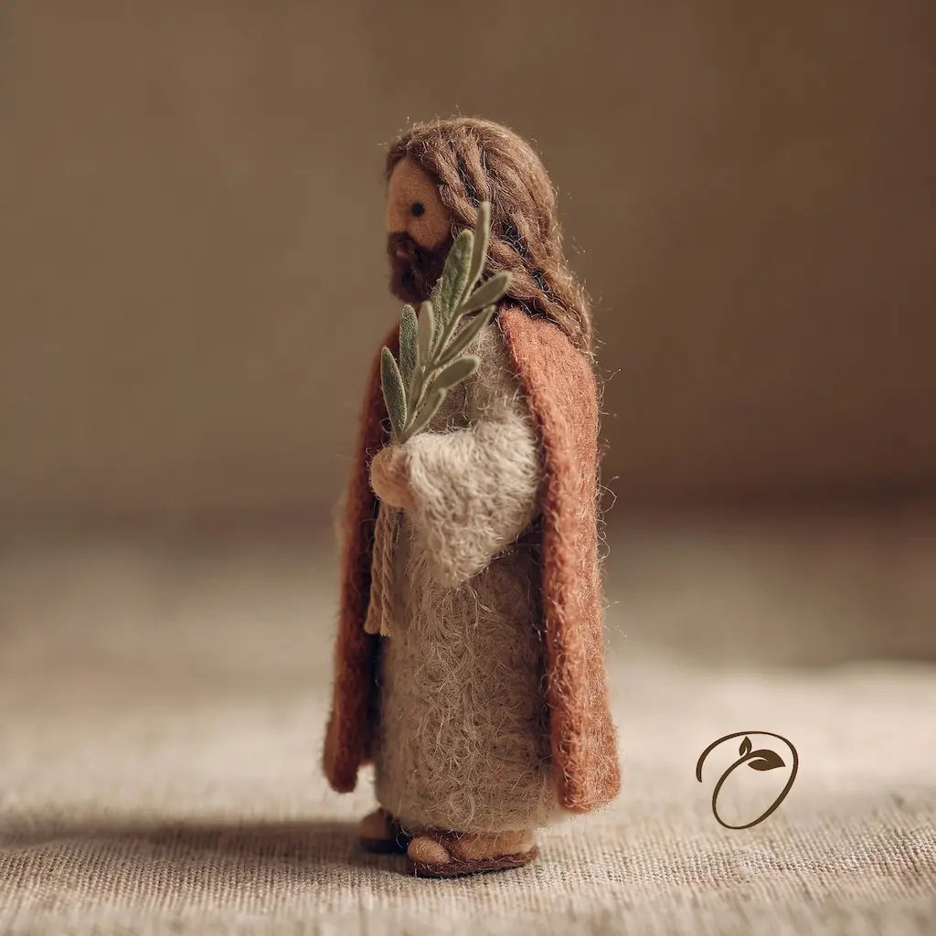 Handmade felted wool figurine of Jesus Christ holding a green olive branch, standing on a textured beige surface against a warm, neutral background.
