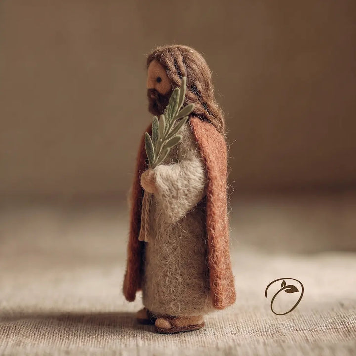 Handmade felted wool figurine of Jesus Christ holding a green olive branch, standing on a textured beige surface against a warm, neutral background.