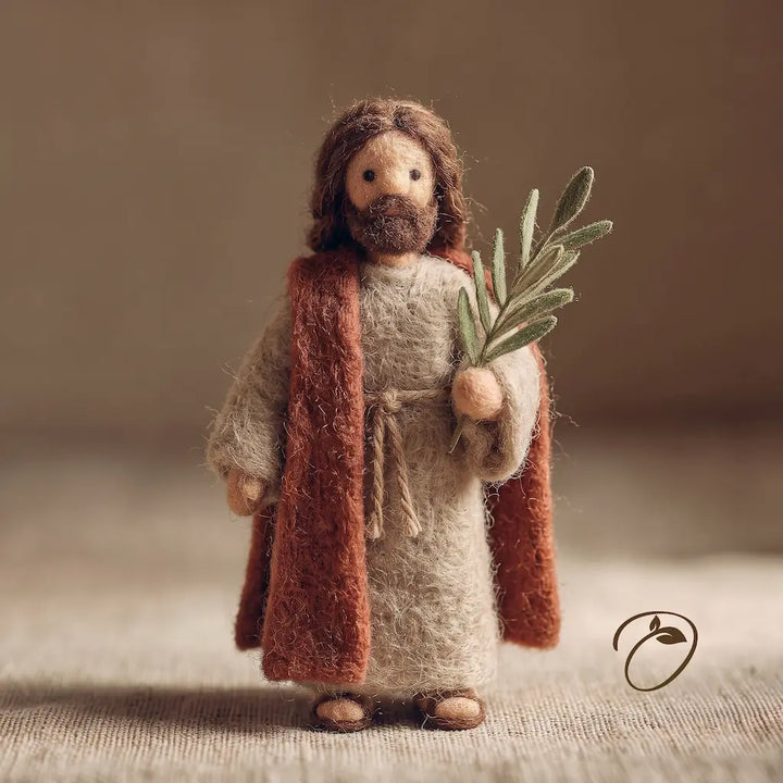 A handcrafted felted wool figurine of Jesus Christ with brown hair and beard, wearing a beige robe and a rust-brown cloak, holding a green olive branch, set against a warm neutral background.